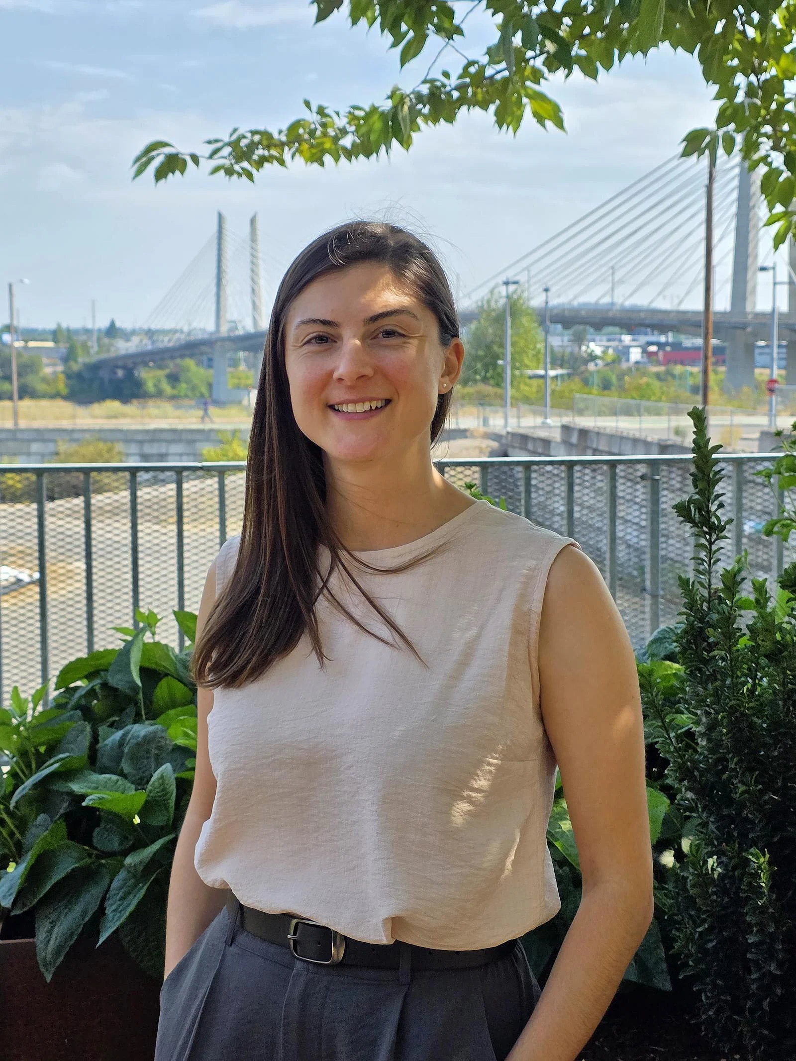 Young female post-doctoral researcher Cigdem Ak stands outside in front of plants, with bridge in background and blue sky.