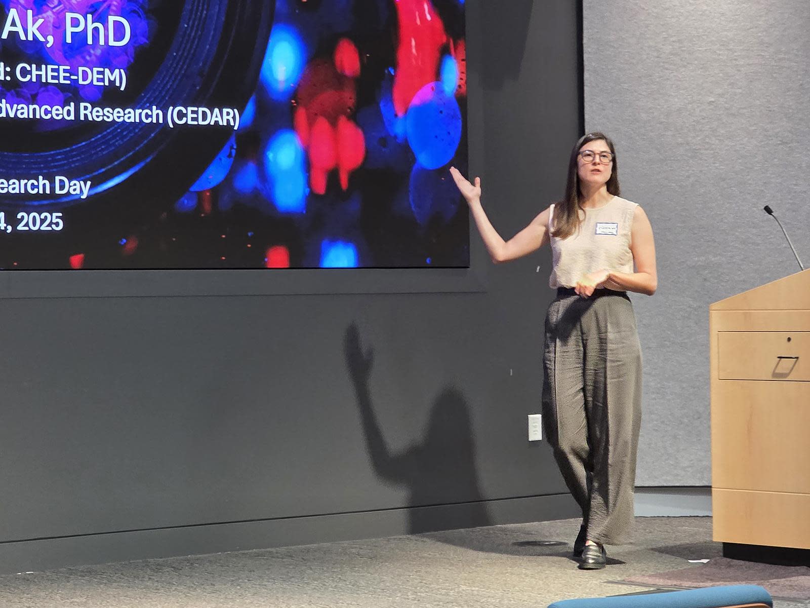 A young woman named Cigdem Ak, a post-doctoral researcher at OHSU, stands on stage next to a podium, giving a presentation on her research project, gesturing to the large screen behind her.